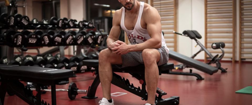Strong, determined man in white tank top sitting in gym, showcasing fitness and strength.